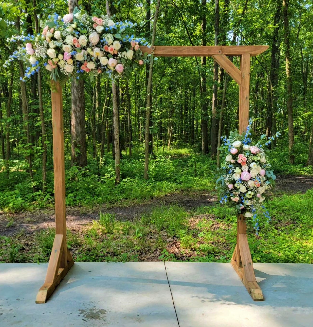 A square arch in front of a forest of trees with white draping and installments of pink and white flowers